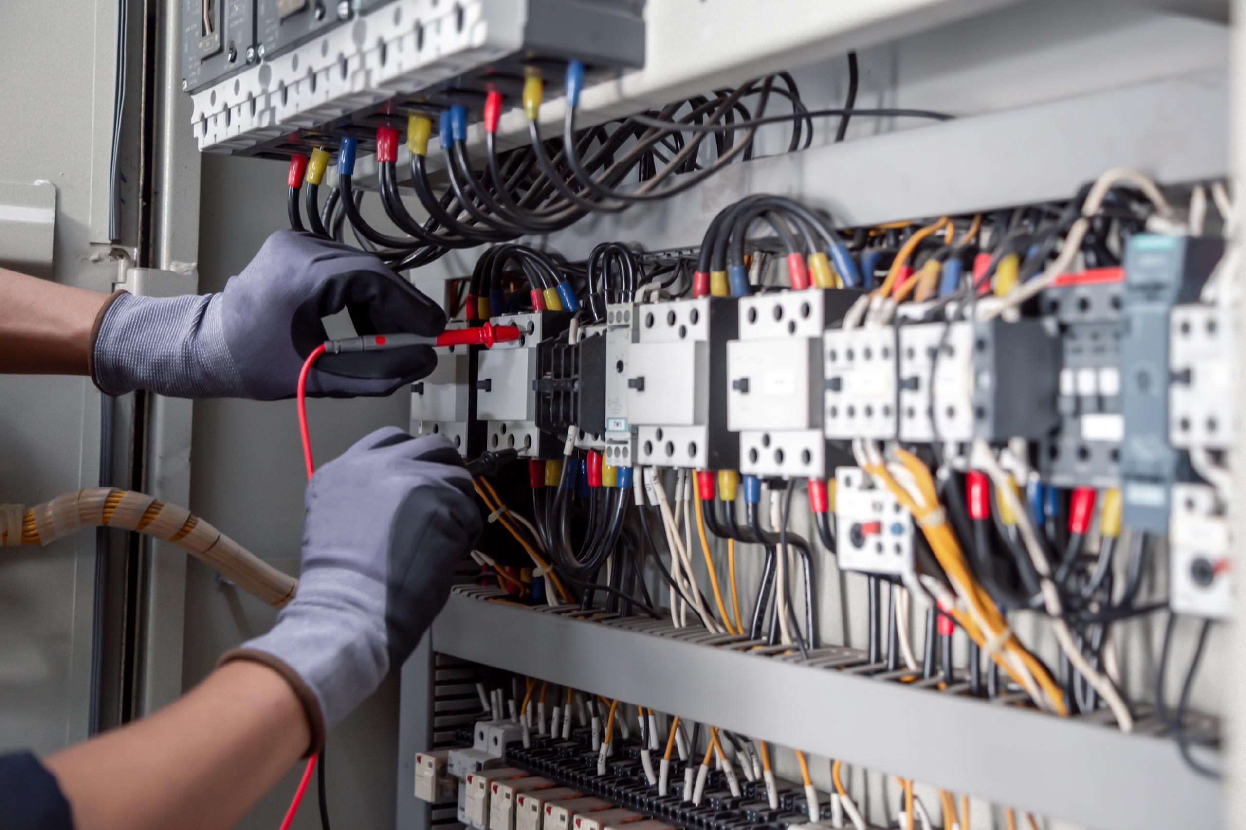 Electrician testing wires in control panel with multimeter inside industrial facility.