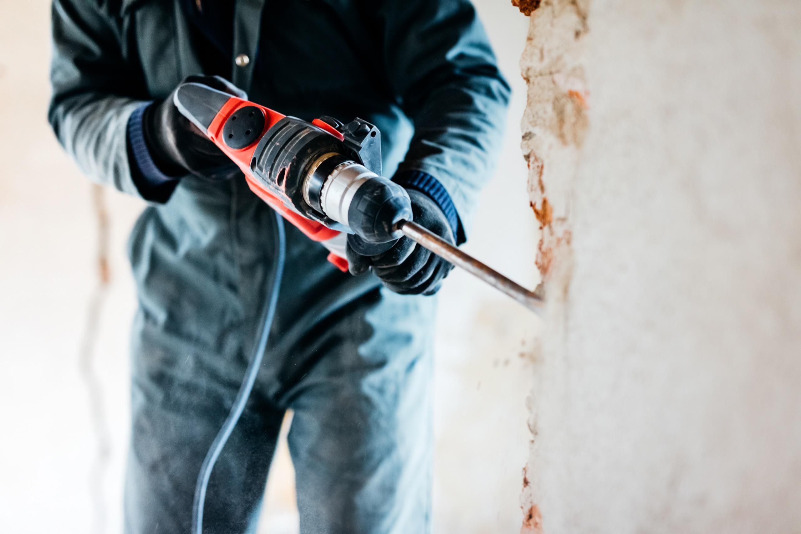 Worker wearing gloves and coveralls using a power drill on a wall during renovation work.