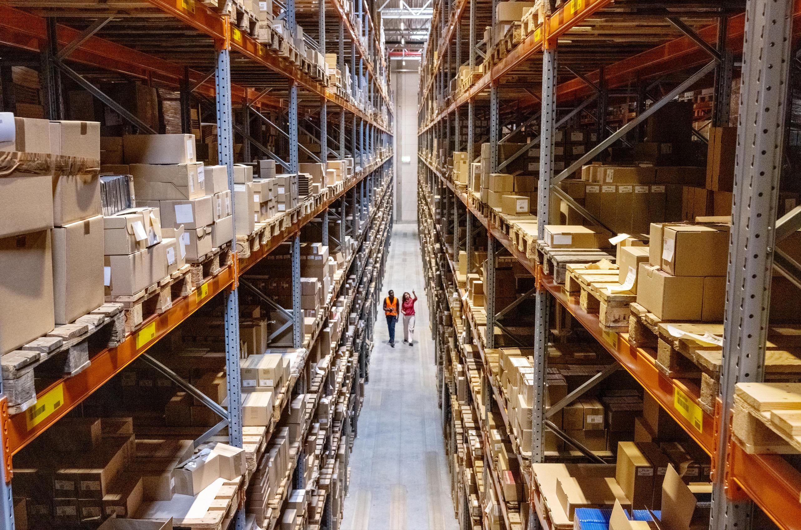 Workers walking inside large warehouse with high racks full of boxes.