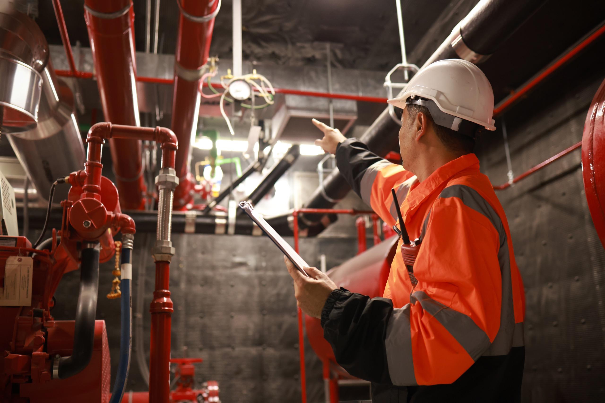 Engineer in high-visibility jacket pointing and inspecting red mechanical pipes and fire safety systems.
