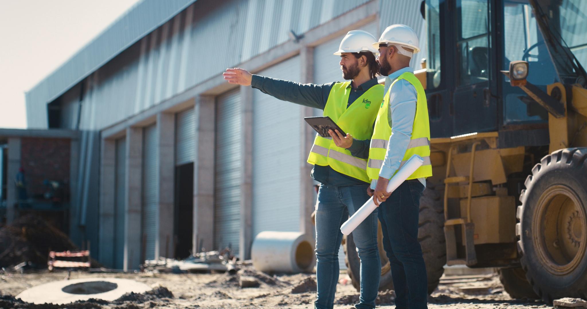 Two construction managers in safety gear reviewing plans and pointing across an active worksite with heavy machinery in the background.