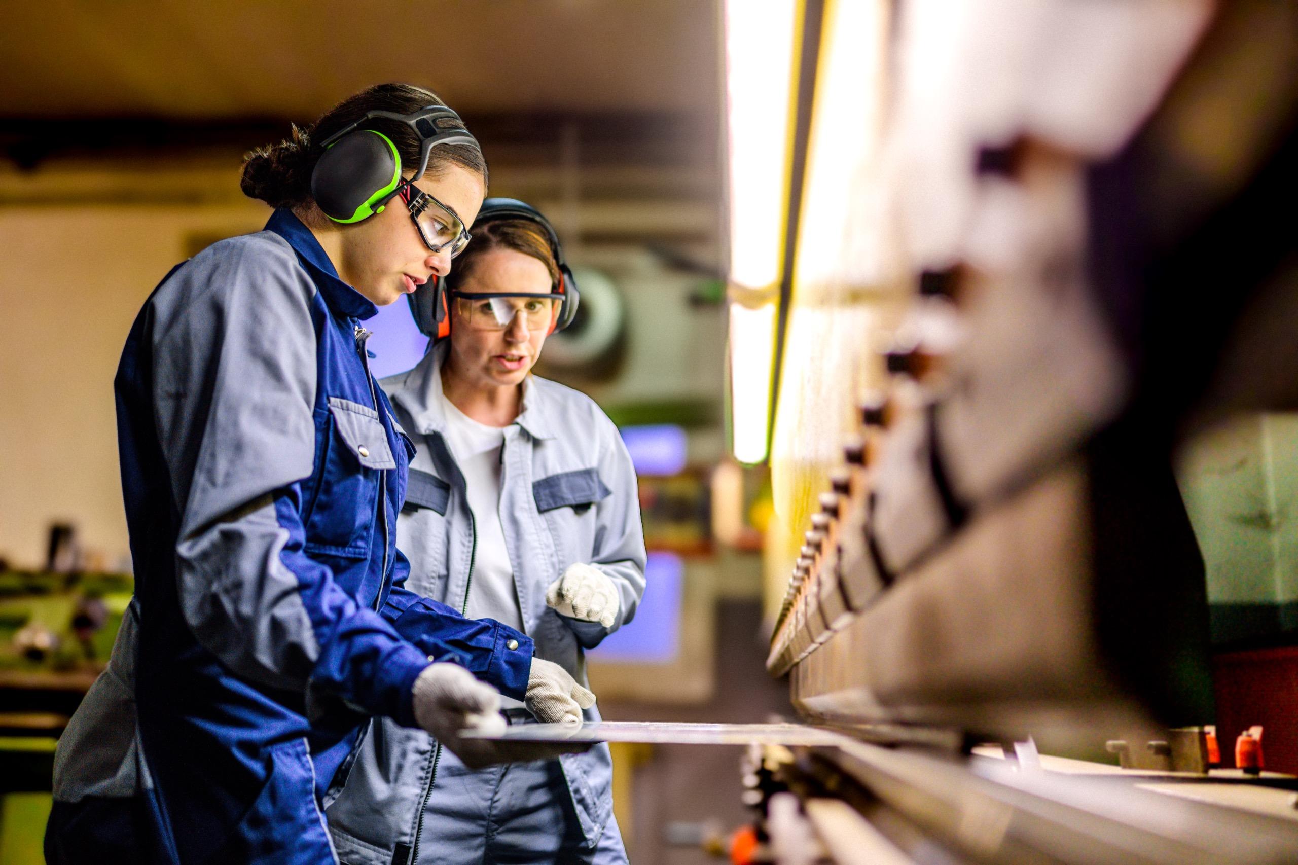 Two technicians in safety goggles and ear protection working together to operate industrial machinery in a manufacturing facility.