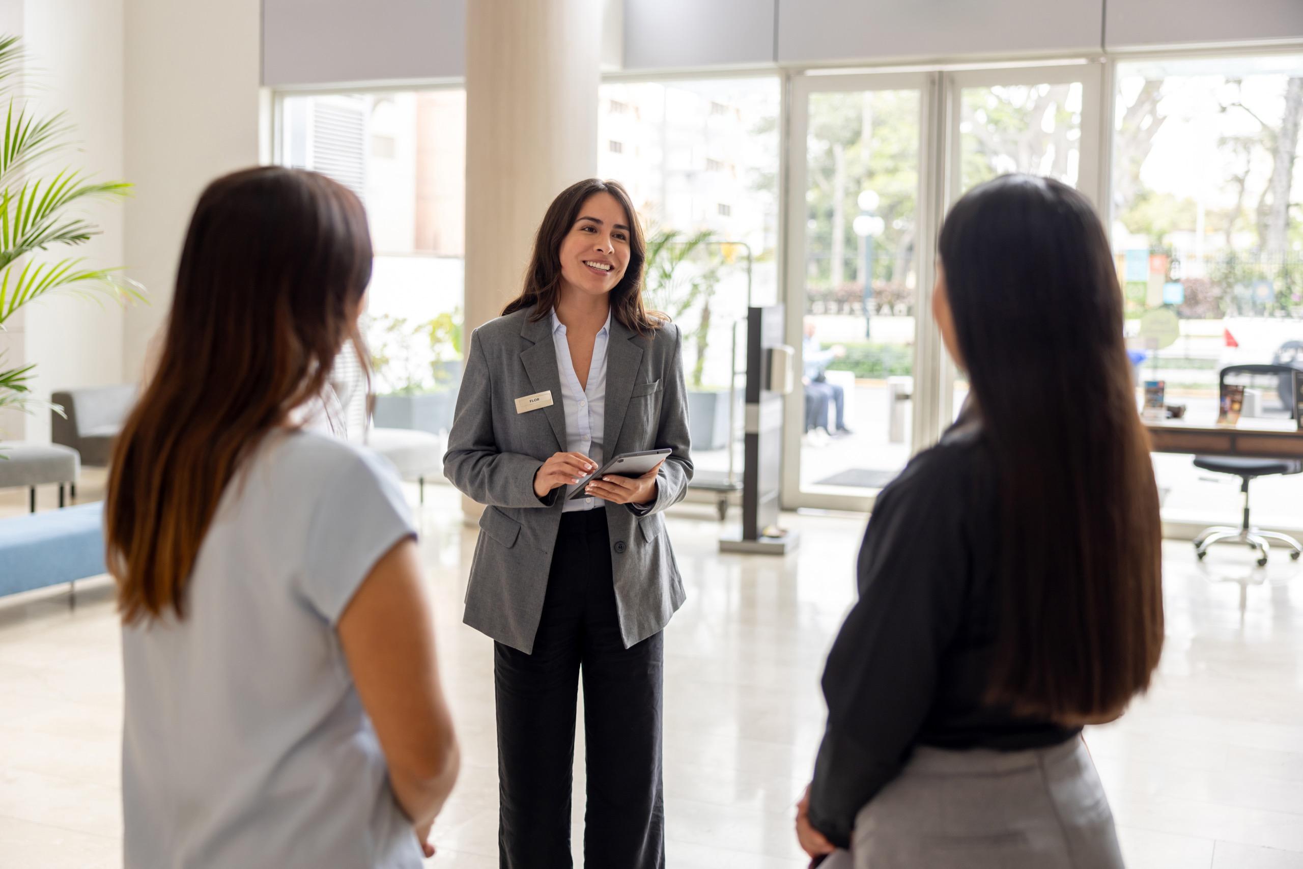 Smiling concierge speaking with guests while holding a tablet in a modern, light-filled lobby.