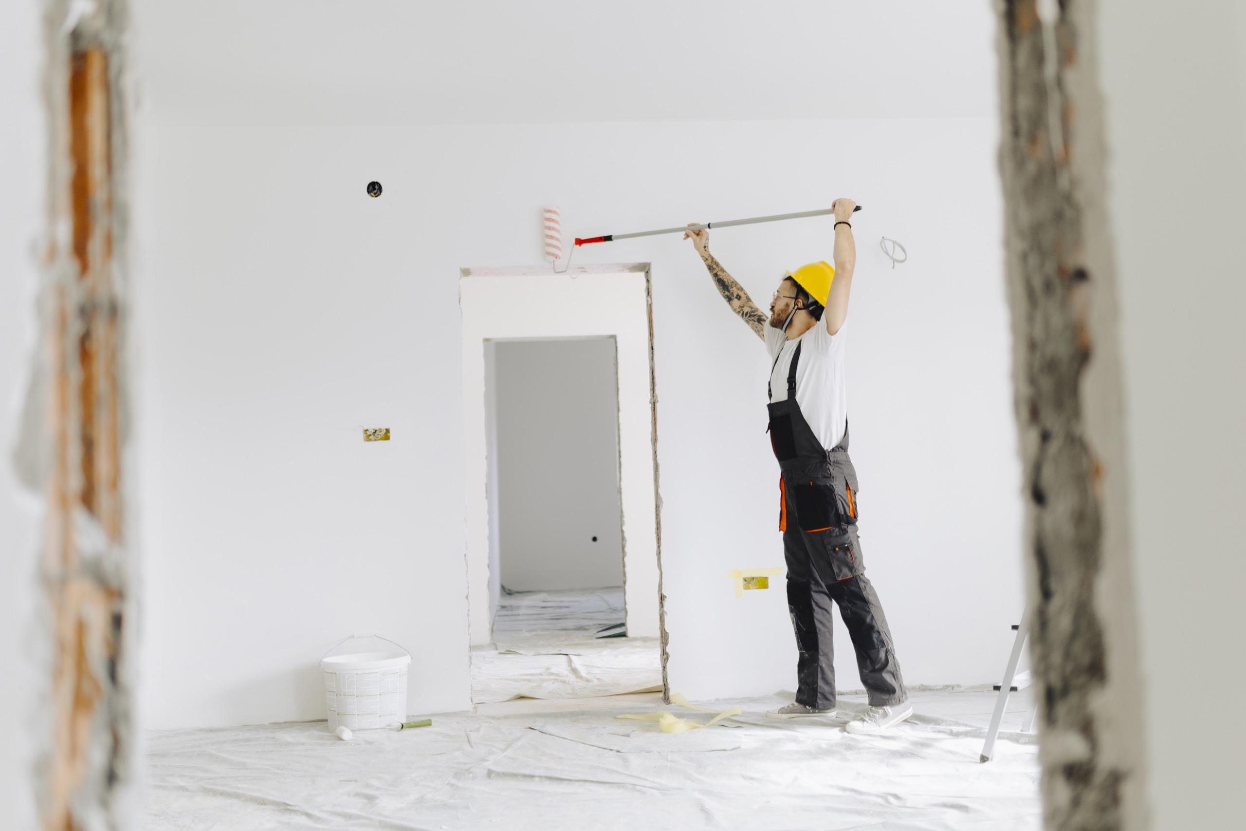 Worker in protective gear using roller to paint interior wall during building renovation.